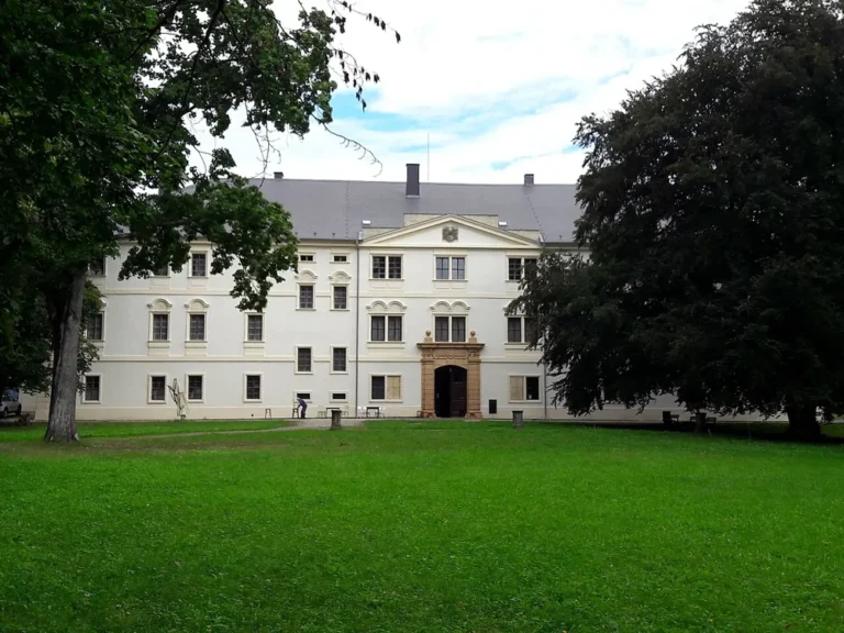 Světlá hlavní budova zámku Křinec za trávníkem, rámovaná korunami stromů